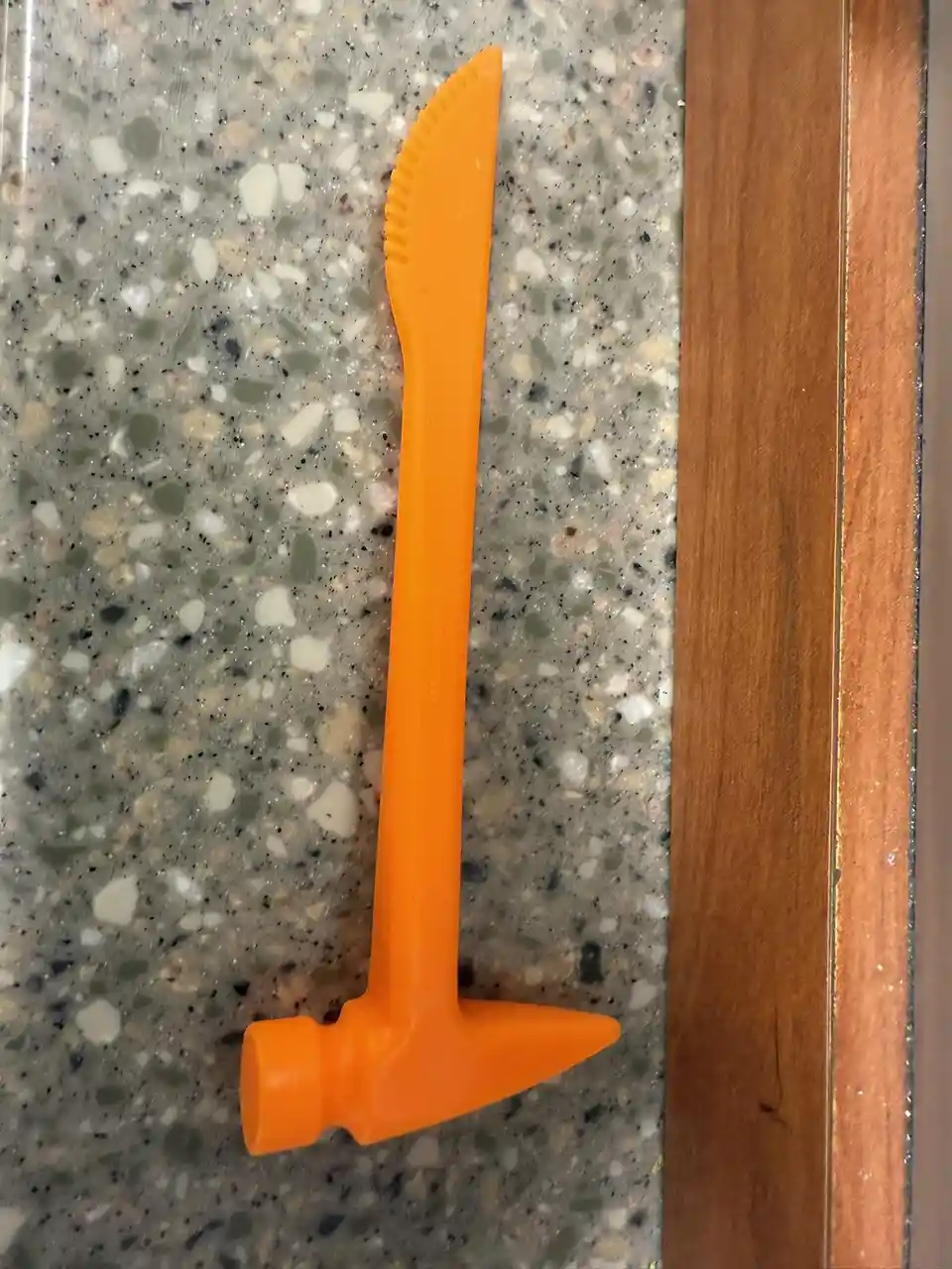 An orange plastic tool with a hammer-like handle and a serrated blade tip rests on a speckled gray countertop next to a…