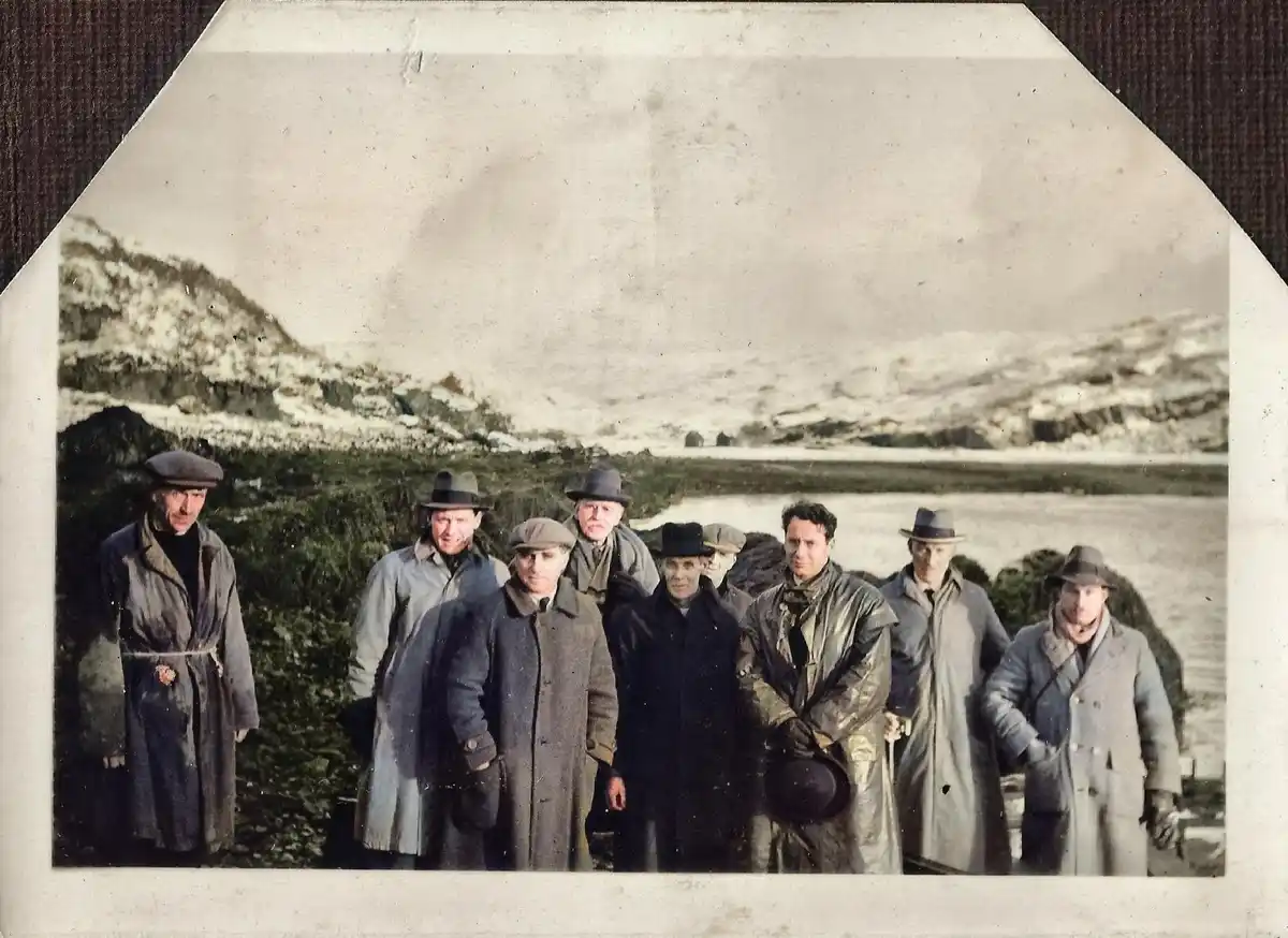 A vintage, sepia-toned photograph shows a group of nine men in heavy coats and hats standing outdoors near a body of water…
