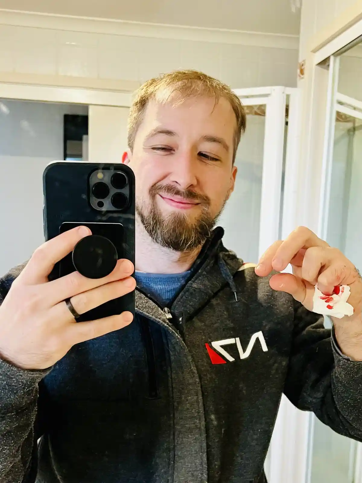 A man with a beard and short hair takes a mirror selfie holding a smartphone with a pop socket, smiling smugly while showing…