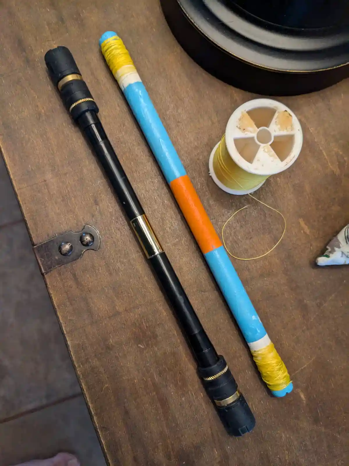 Two objects lie on a wooden surface: one is a black, segmented object resembling a lightsaber hilt, and the other is a…