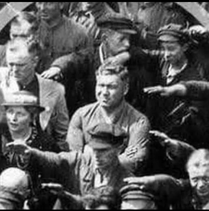 A grainy black-and-white historical photograph shows a man in the center of a crowd, surrounded by people reaching toward…