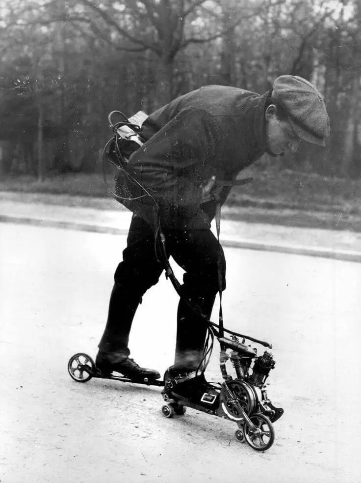 A black-and-white historical photo shows a man in early 20th-century clothing, including a flat cap and boots, bent over…