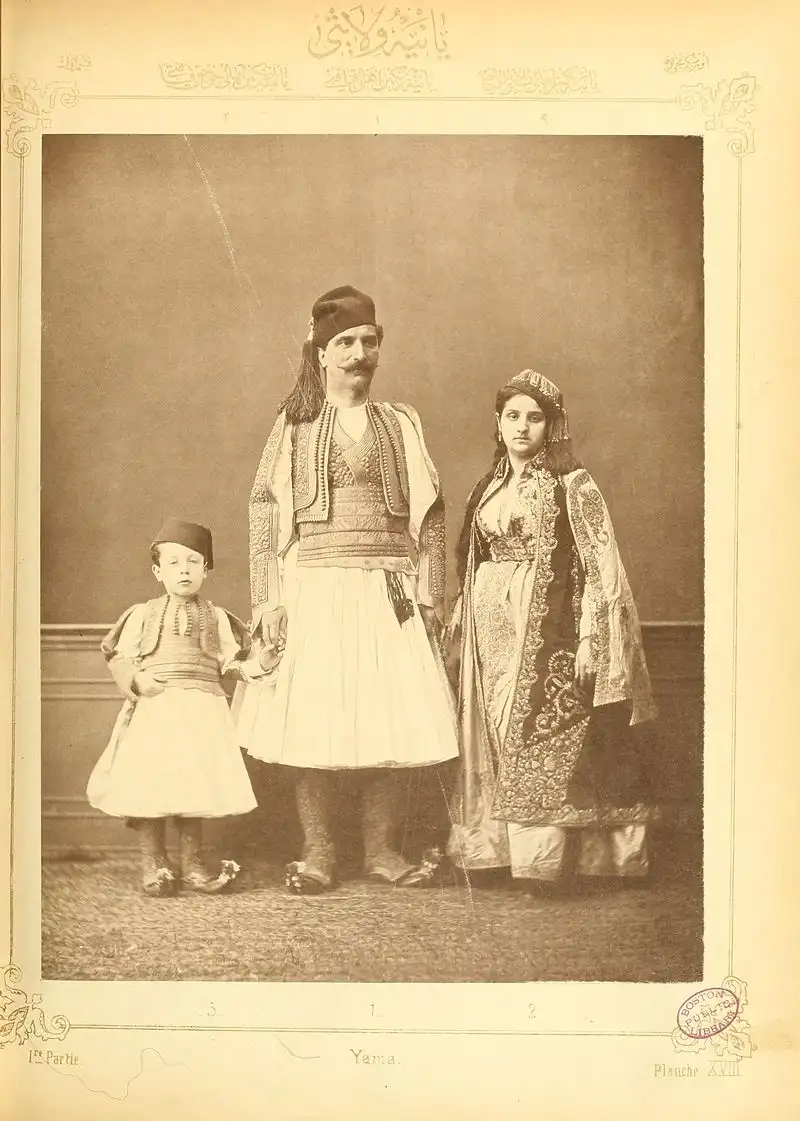 A sepia-toned vintage photograph shows a man, woman, and young boy dressed in ornate traditional attire, standing formally…