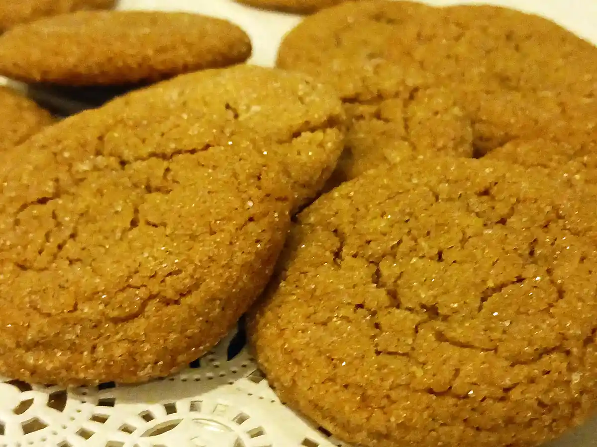 A close-up photo of several golden-brown, textured cookies with visible sugar crystals, resting on a white decorative doily…
