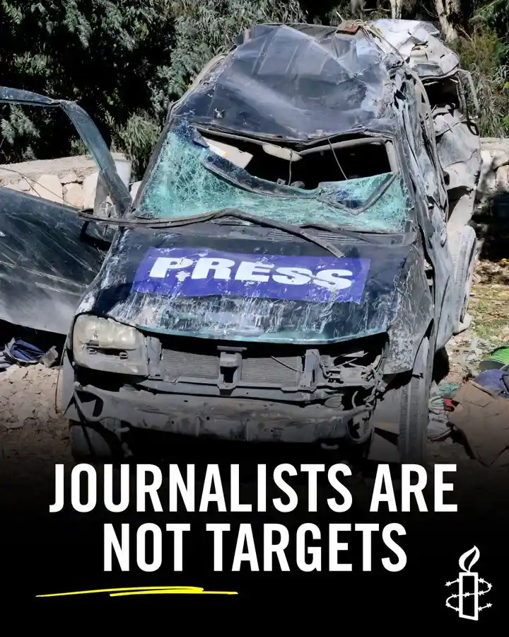 A heavily damaged black vehicle with a 'PRESS' decal on the hood sits in rubble, its windshield shattered and doors bent…