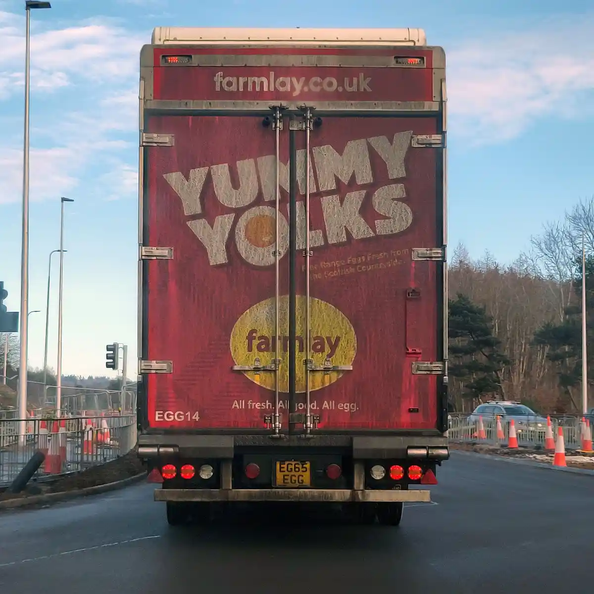 The rear of a red delivery truck with 'farmlay.co.uk' and 'YUMMY YOLKS' branding, featuring a yellow egg logo and slogan…