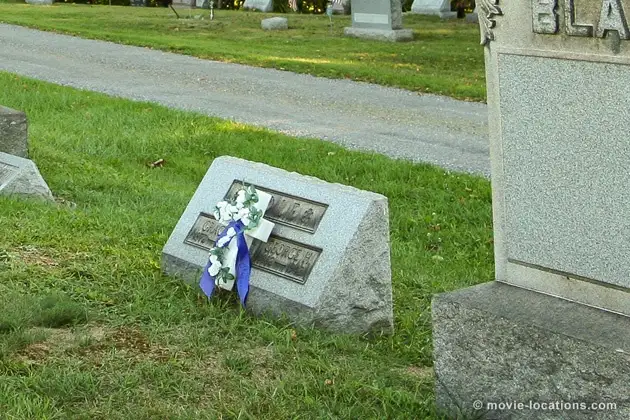 A tilted, fallen gravestone in a cemetery with a blue and white floral wreath tied to it. The stone is partially buried in…