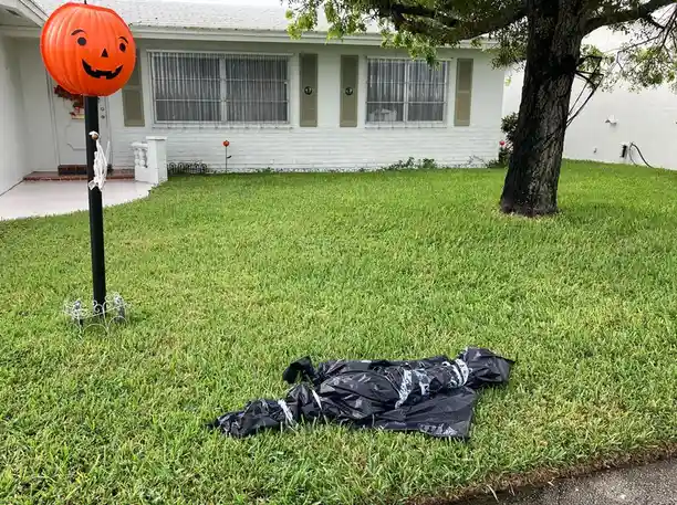 A grassy front yard features a large orange jack-o'-lantern decoration on a pole next to a house, while a black plastic bag…