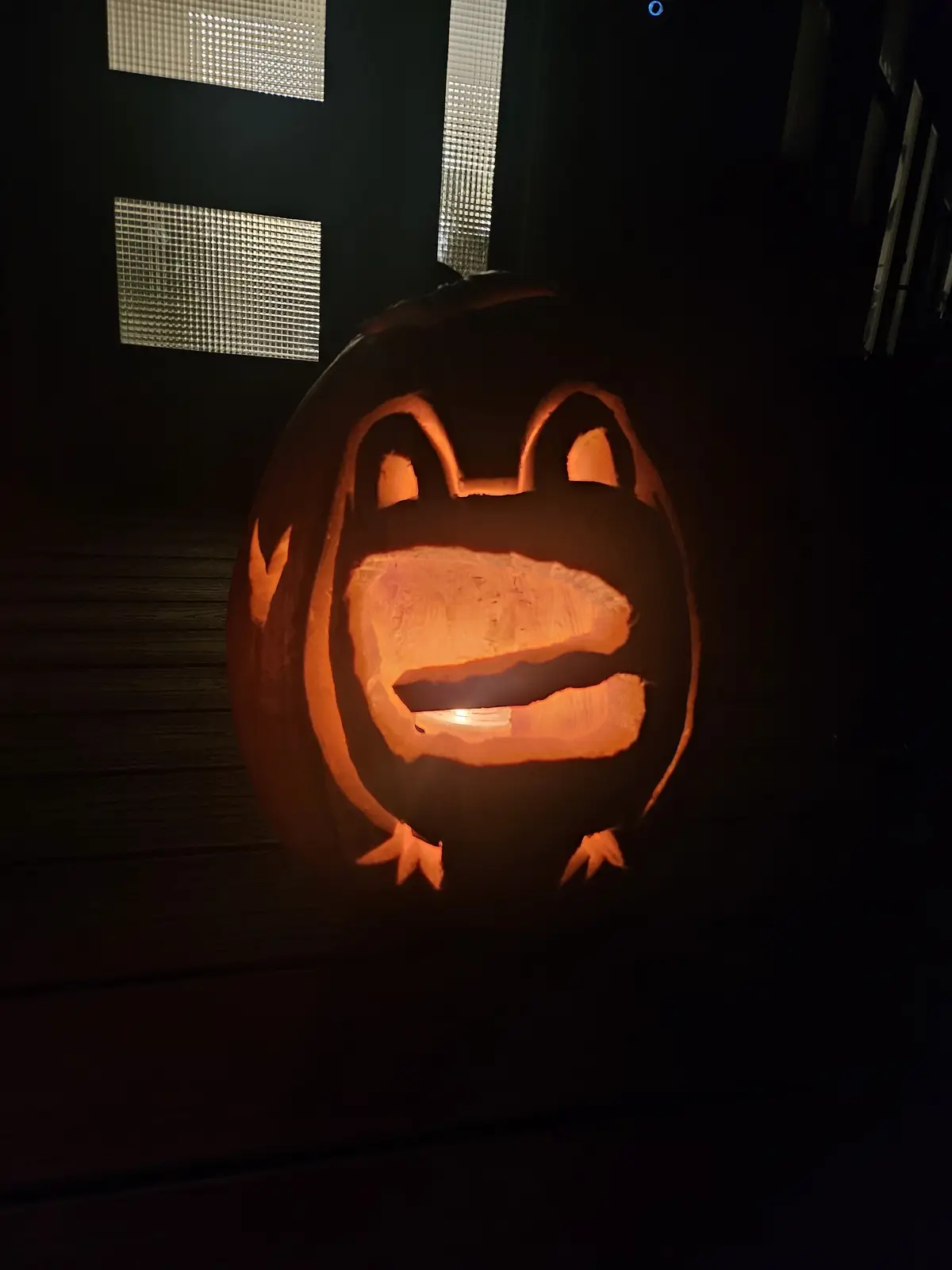 A carved Halloween pumpkin with a wide, open-mouthed frog-like face, illuminated from within, sits on a dark wooden surface…