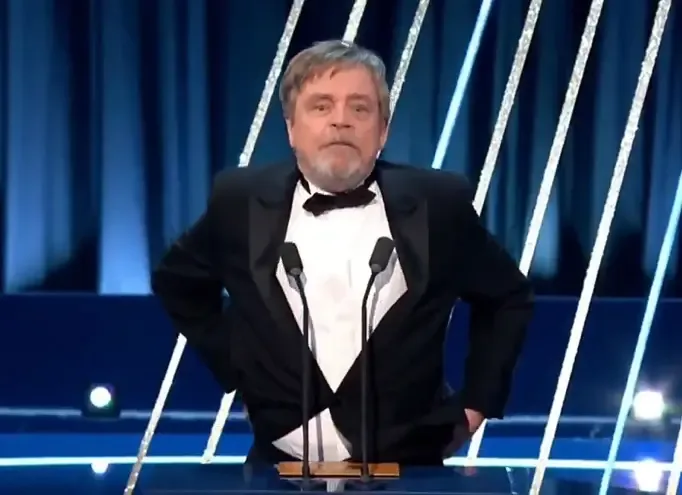 a man in a black tuxedo with a bowtie stands at a podium with two microphones, hands on hips, looking displeased. Behind him…