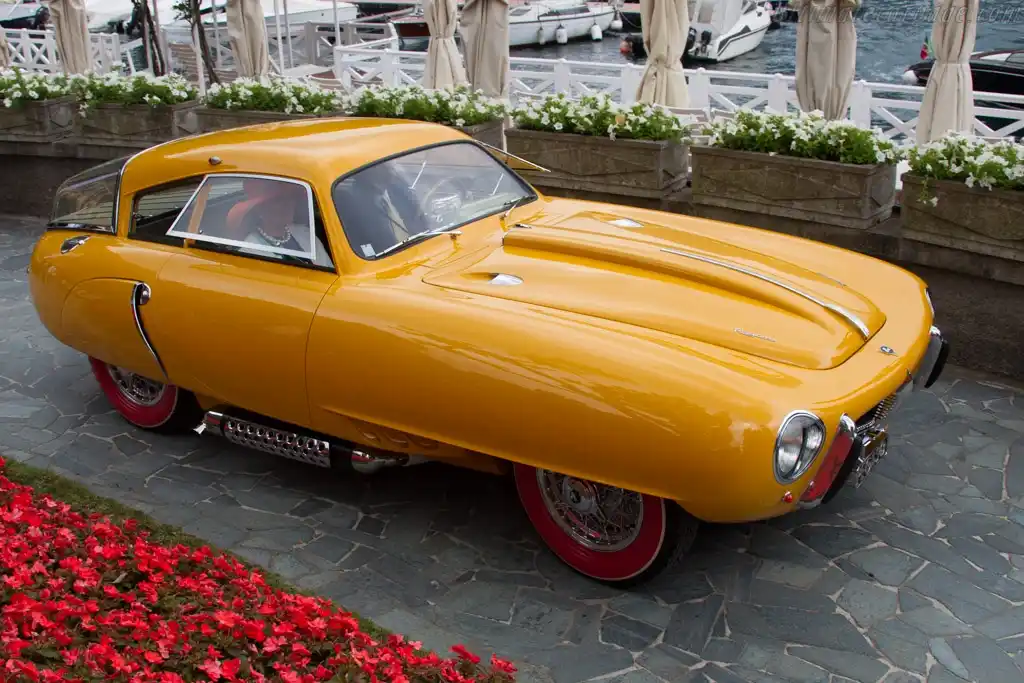A bright yellow vintage sports car with a sleek, aerodynamic design and red-rimmed wheels is parked on a stone-paved area…