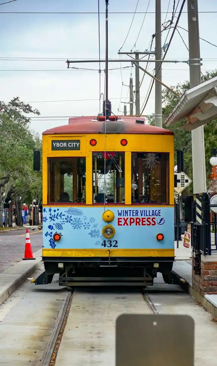 A yellow and red trolley car, labeled 'YBOR CITY' at the top and 'WINTER VILLAGE EXPRESS' on the front with decorative blue…