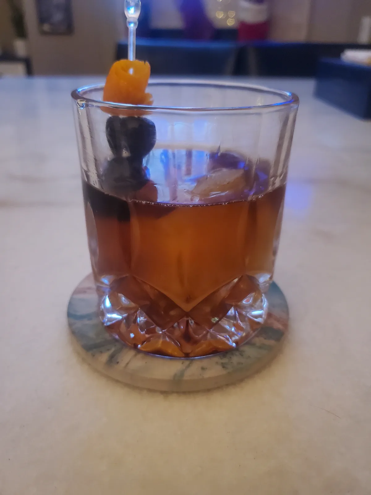 A close-up photo of a cocktail in a faceted rocks glass, resting on a marble coaster. The drink is amber-colored with ice…
