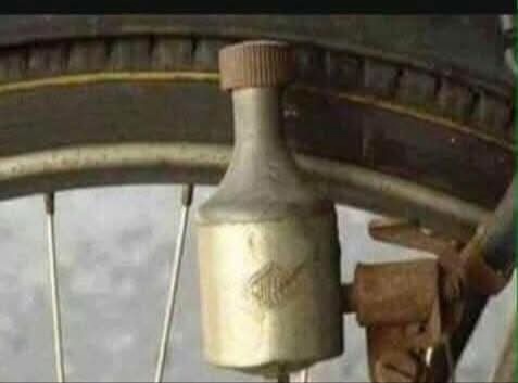 a close-up of a vintage bicycle wheel with a unique, bottle-shaped metal bell attached to the spokes. The bell has a cap on…