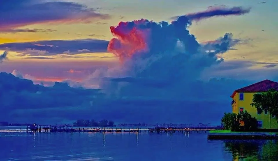 A scenic sunset or sunrise over water with a large, colorful cloud formation dominating the sky. A yellow building with a…