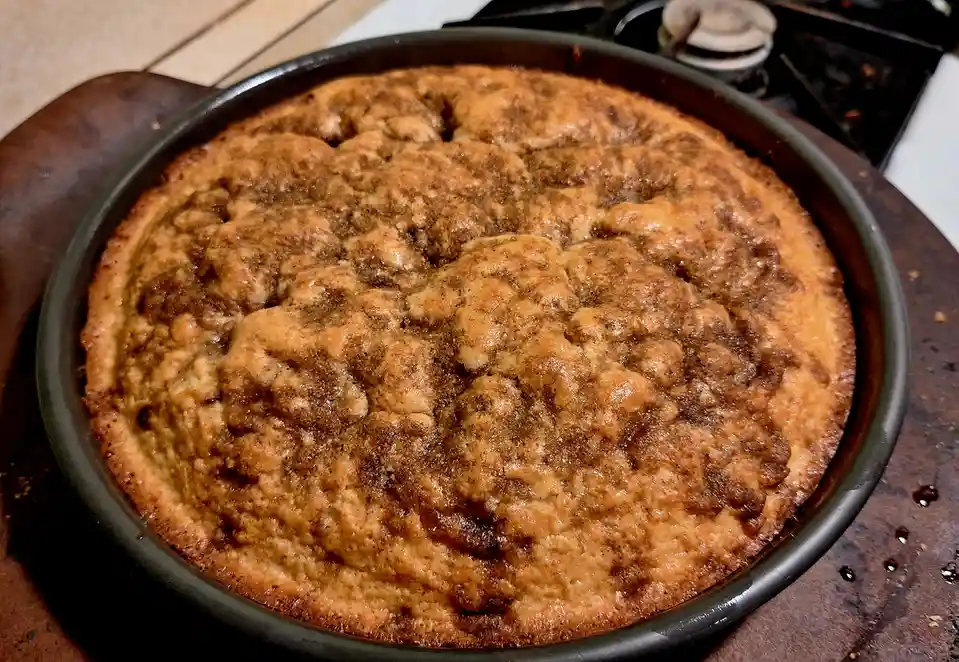 A close-up photo shows a round baked dessert in a dark metal pan, resting on a wooden surface. The cake has a golden-brown…