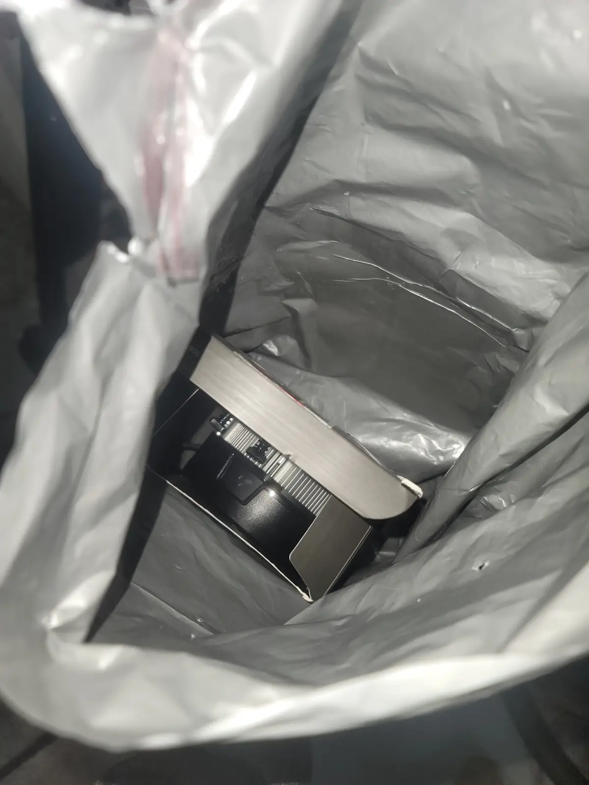 A close-up shot shows a vintage-style black and silver camera nestled inside a crumpled, translucent plastic trash bag. The…