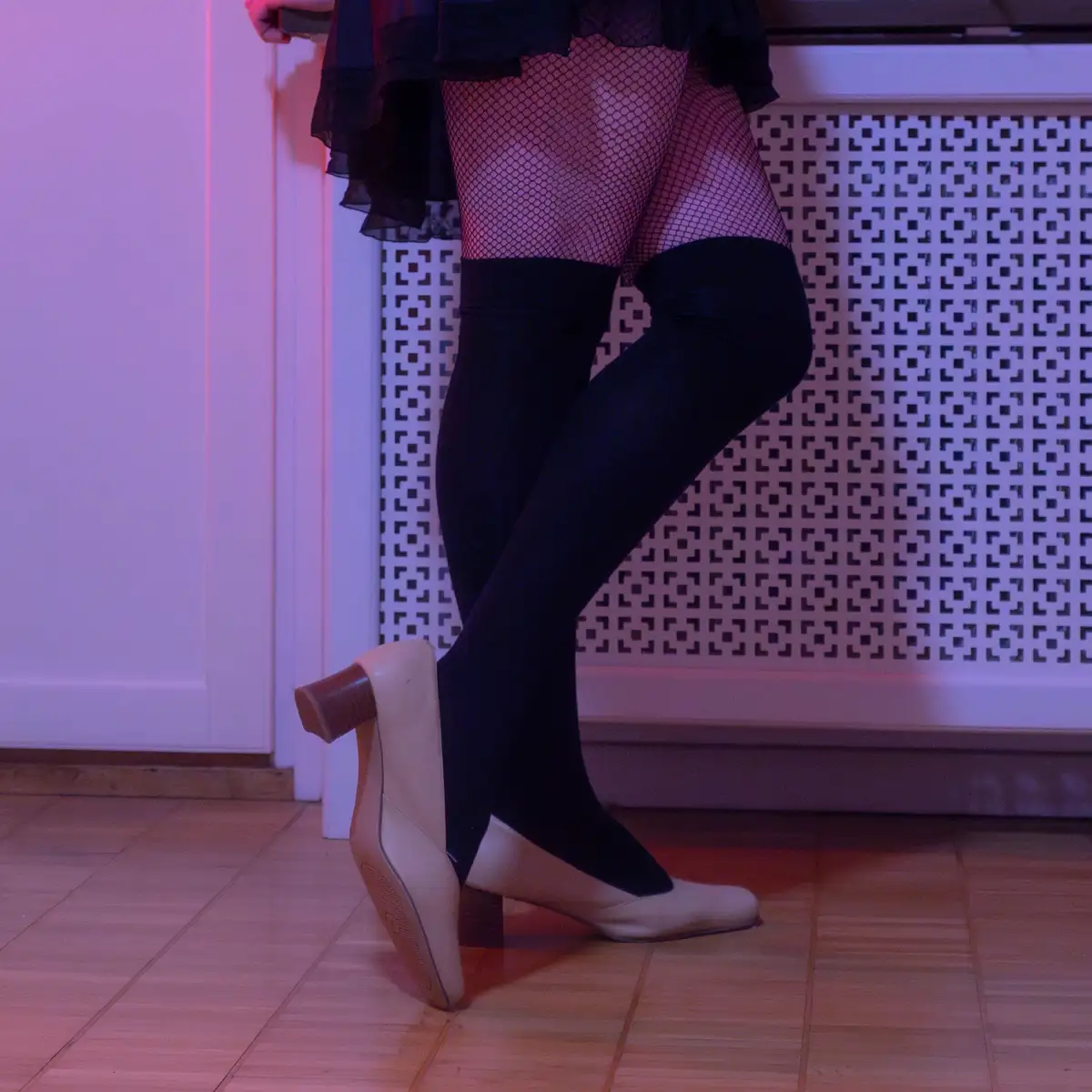 A person wearing black fishnet stockings and black thigh-high socks stands on a tiled floor, one foot in a beige heel shoe…