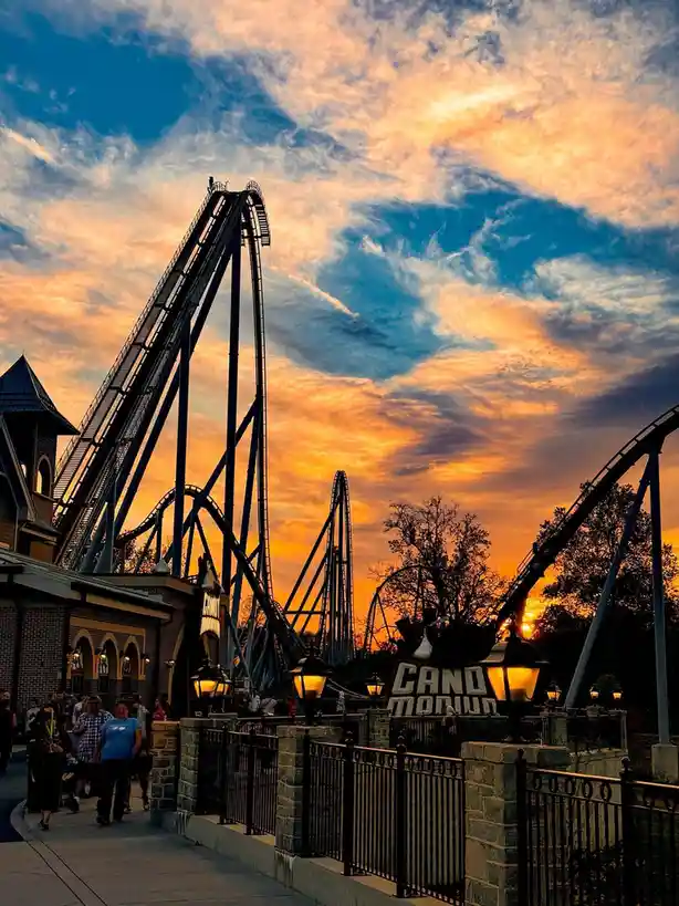 A scenic photo of a roller coaster at an amusement park during sunset, with vibrant orange and blue clouds in the sky. The…