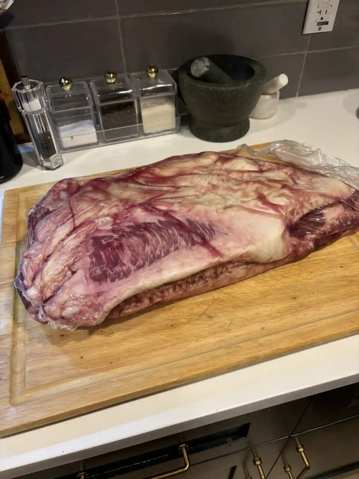 A large, raw, whole beef carcass is wrapped in plastic and sits on a wooden cutting board on a kitchen counter. In the…