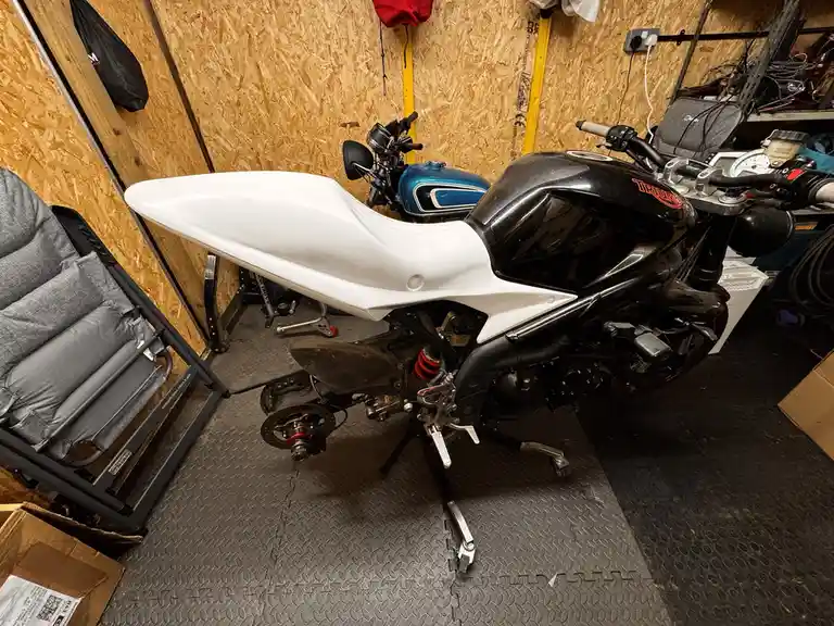 A black Triumph motorcycle with a white seat is shown in a cluttered garage, propped up on its kickstand. The bike appears…