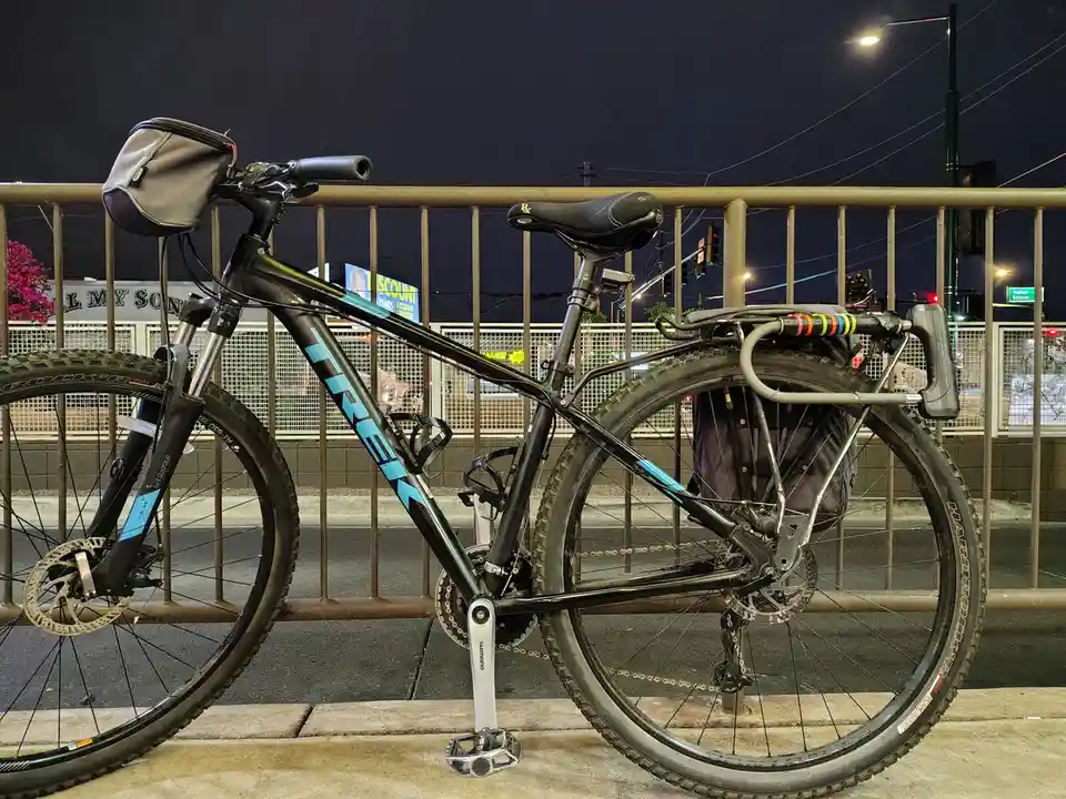 A black Trek mountain bike with a front basket and rear rack is parked at night against a metal railing. The bike has thick…