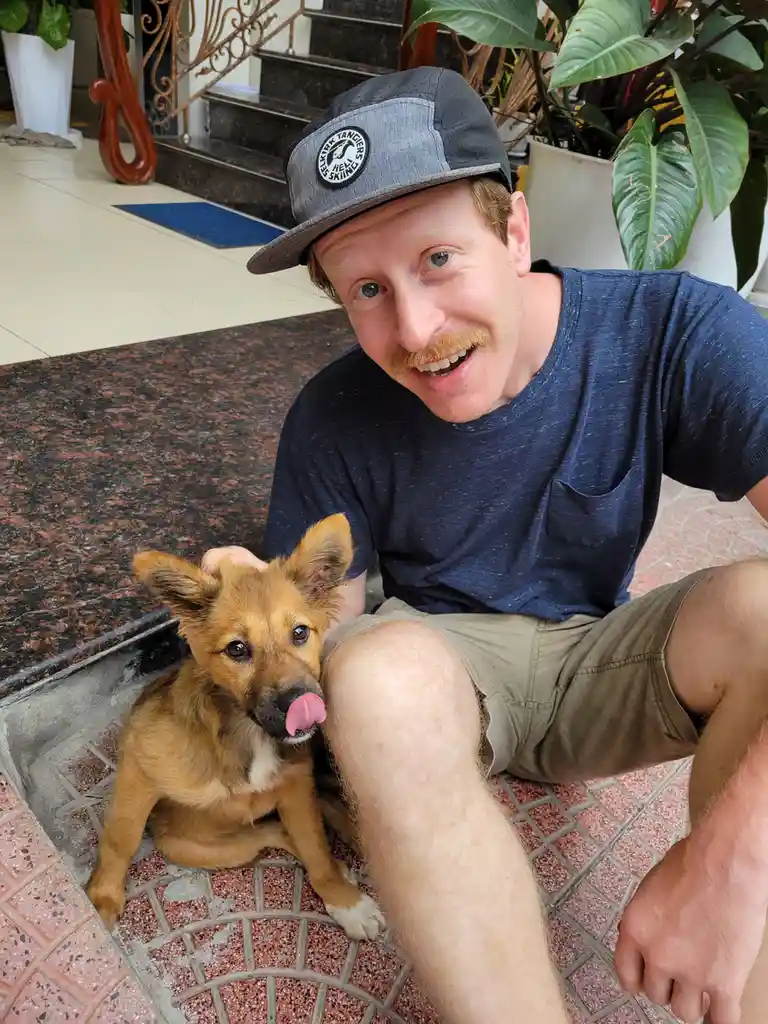 A man with a mustache and a grey baseball cap is crouching on a tiled floor next to a small brown puppy. The puppy is…