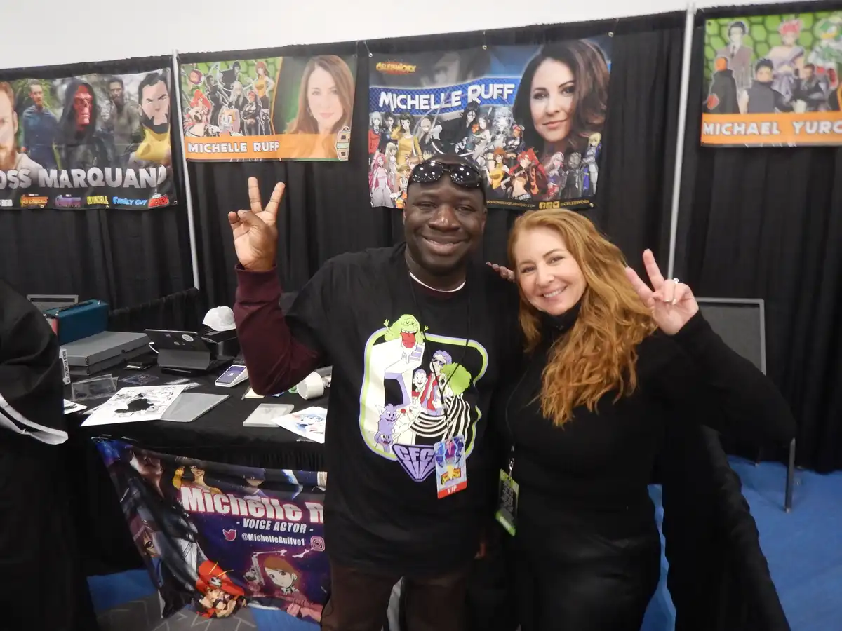 A man and a woman pose together at a convention, both making peace signs. Behind them are banners for voice actors…