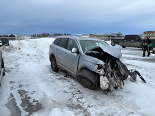 A silver SUV is severely damaged in a snowy parking lot, with its front end crushed and mangled. Two people stand nearby…