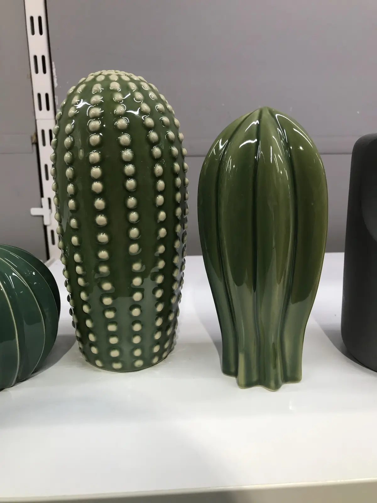Two green ceramic cactus-shaped decorative vases sit on a white shelf, one with raised bead-like bumps and the other with…