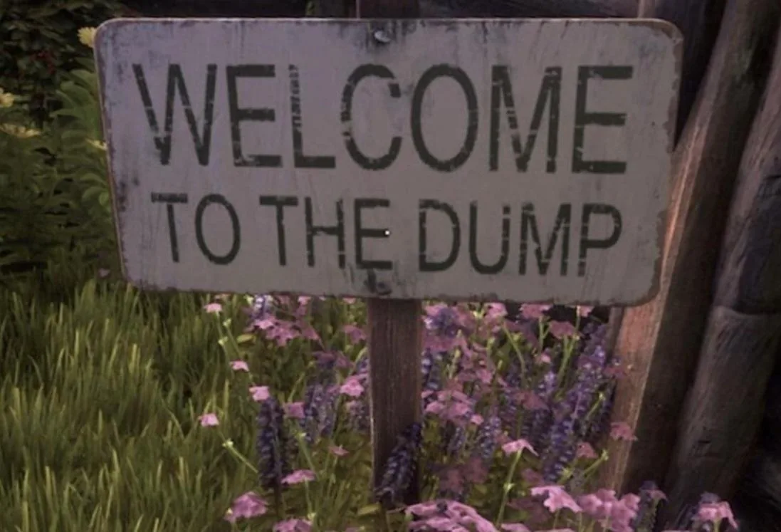 A weathered, white rectangular sign with black stenciled text reading 'WELCOME TO THE DUMP' is mounted on a wooden post in a…