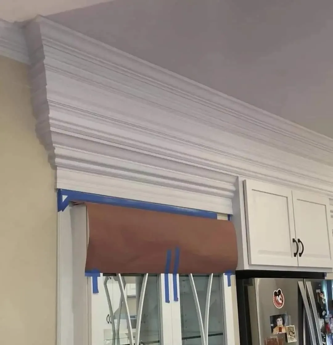 a close-up photo of a kitchen shows a very ornate, multi-layered white crown molding installed above a window and cabinets…