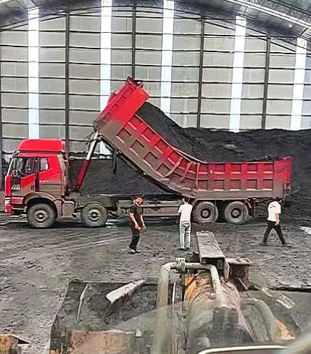 A red dump truck is tipping its bed to unload black coal into a large pile inside a warehouse. Three men in casual clothes…