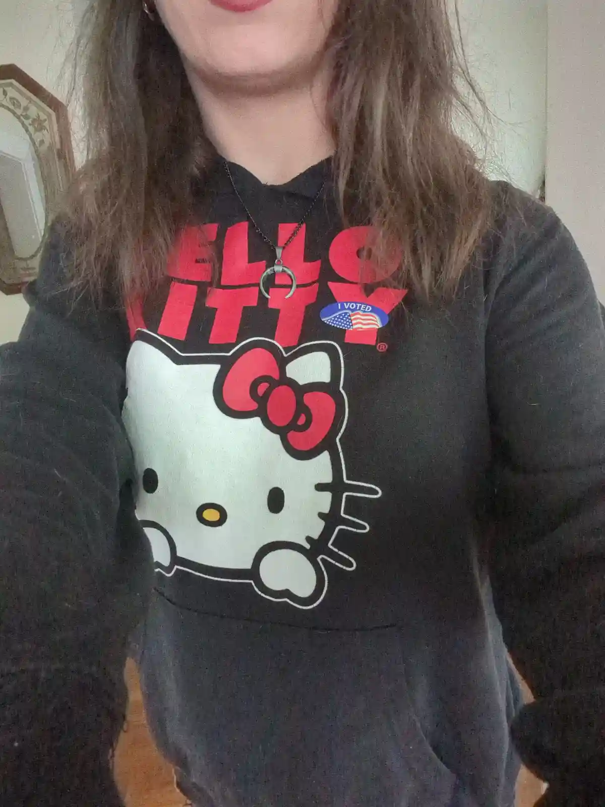 A person wearing a black hoodie with a large Hello Kitty graphic and red text above it, with a small blue 'I VOTED' sticker…