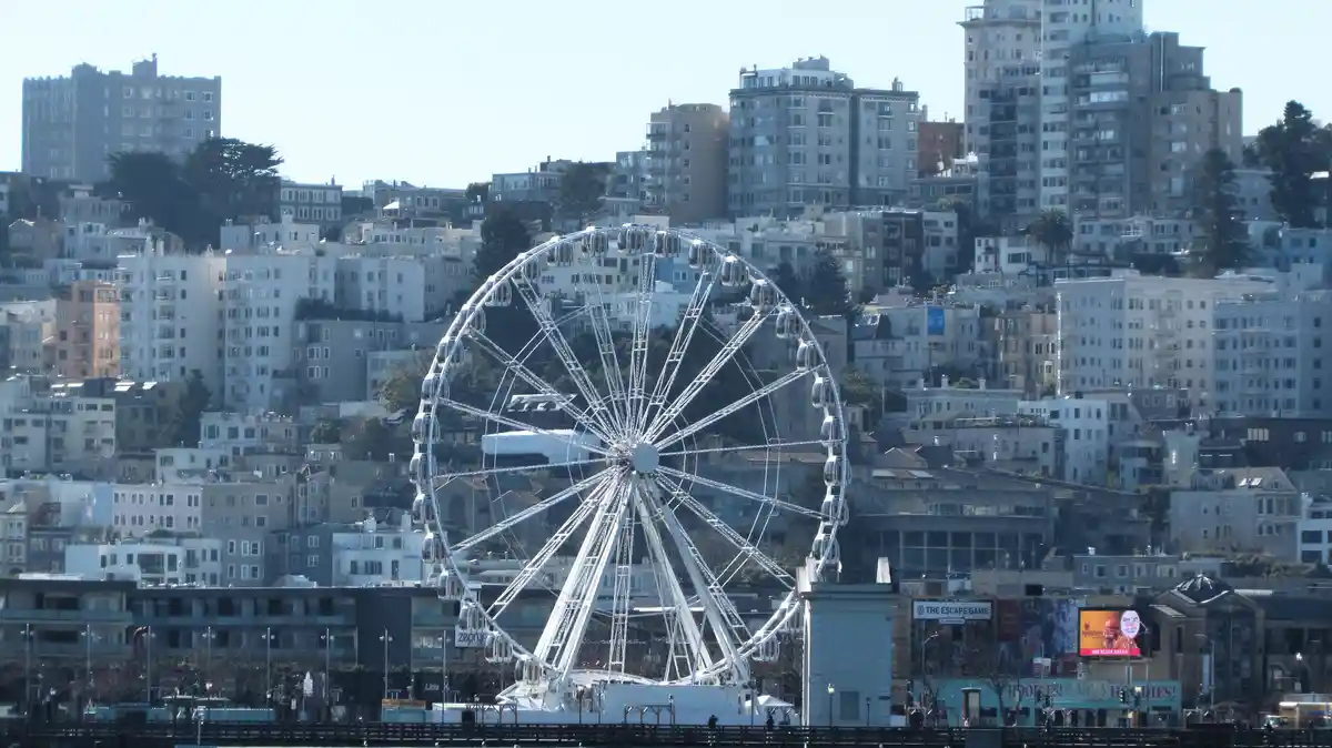 A large white Ferris wheel stands in the foreground against a backdrop of densely packed, multi-story buildings on a…