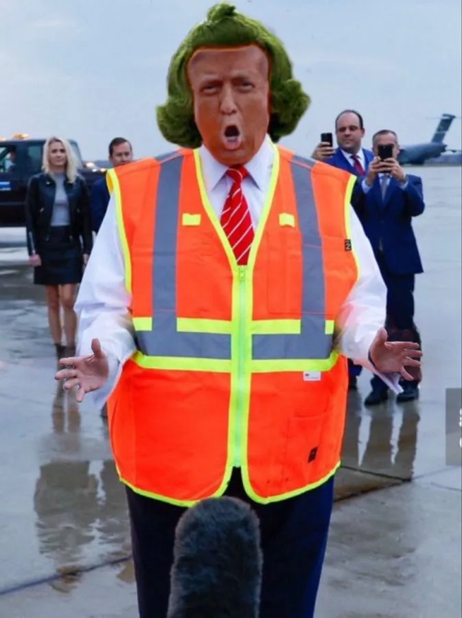 A person wearing a large, exaggerated mask of Donald Trump with green hair, an orange construction safety vest over a white…