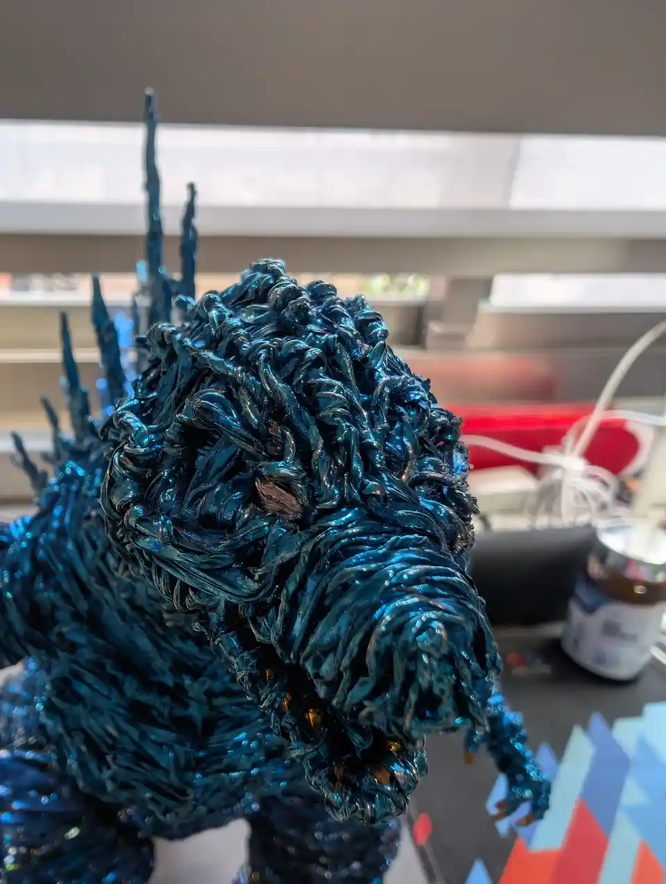 A close-up photo of a blue, textured Godzilla figurine with spiky dorsal fins, positioned on a desk with blurred office…
