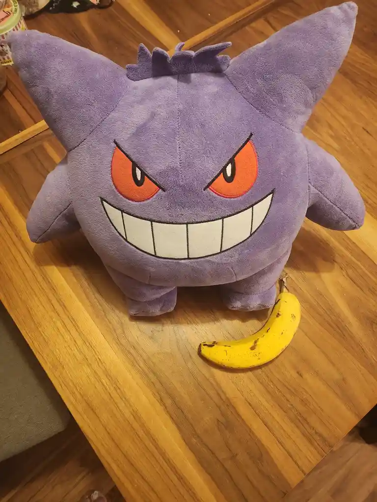 A plush Gengar toy with a menacing grin and red eyes sits on a wooden table, with a ripe yellow banana placed directly in…