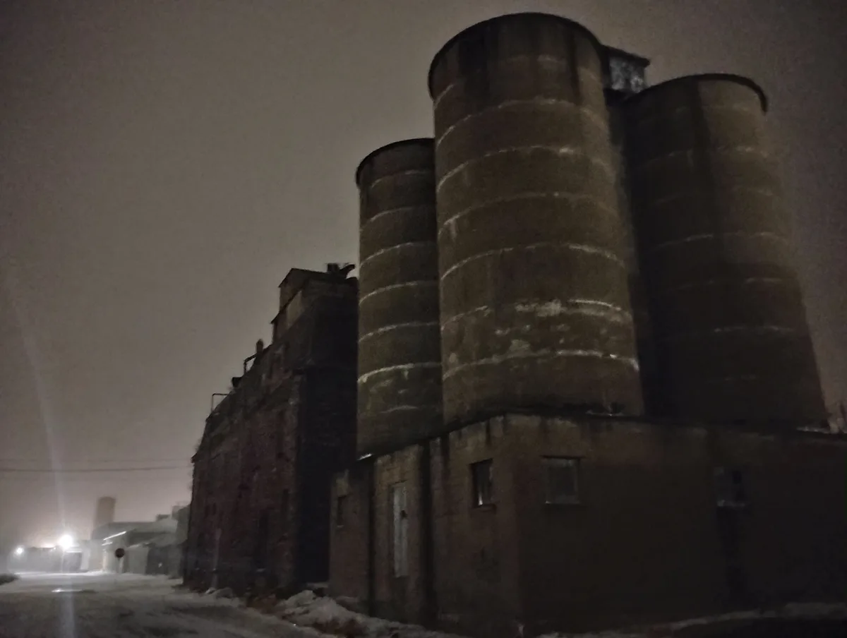 A low-angle night photo shows three large, cylindrical grain silos attached to a brick industrial building, with snow on the…