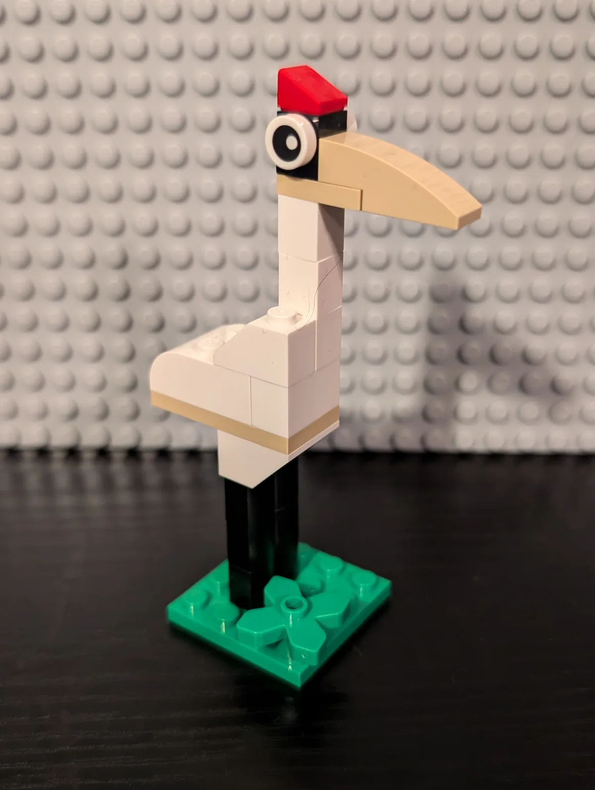 A LEGO bird figure made from white, tan, black, and red pieces stands on a green baseplate against a gray LEGO stud…