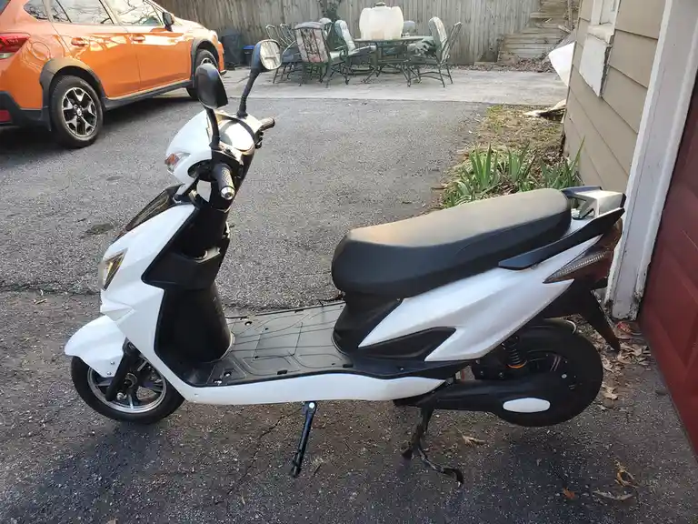 A white and black scooter is parked on an asphalt driveway next to a house, with an orange car visible in the background…