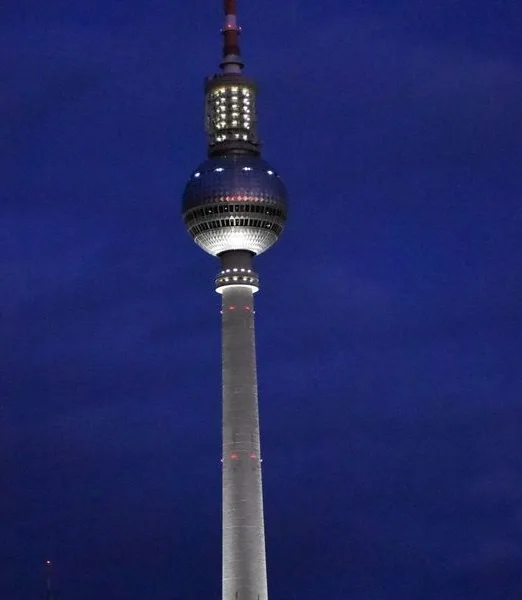 A nighttime photo of the Berlin TV Tower, its illuminated sphere and antenna visible against a dark blue sky. Overlaid text…
