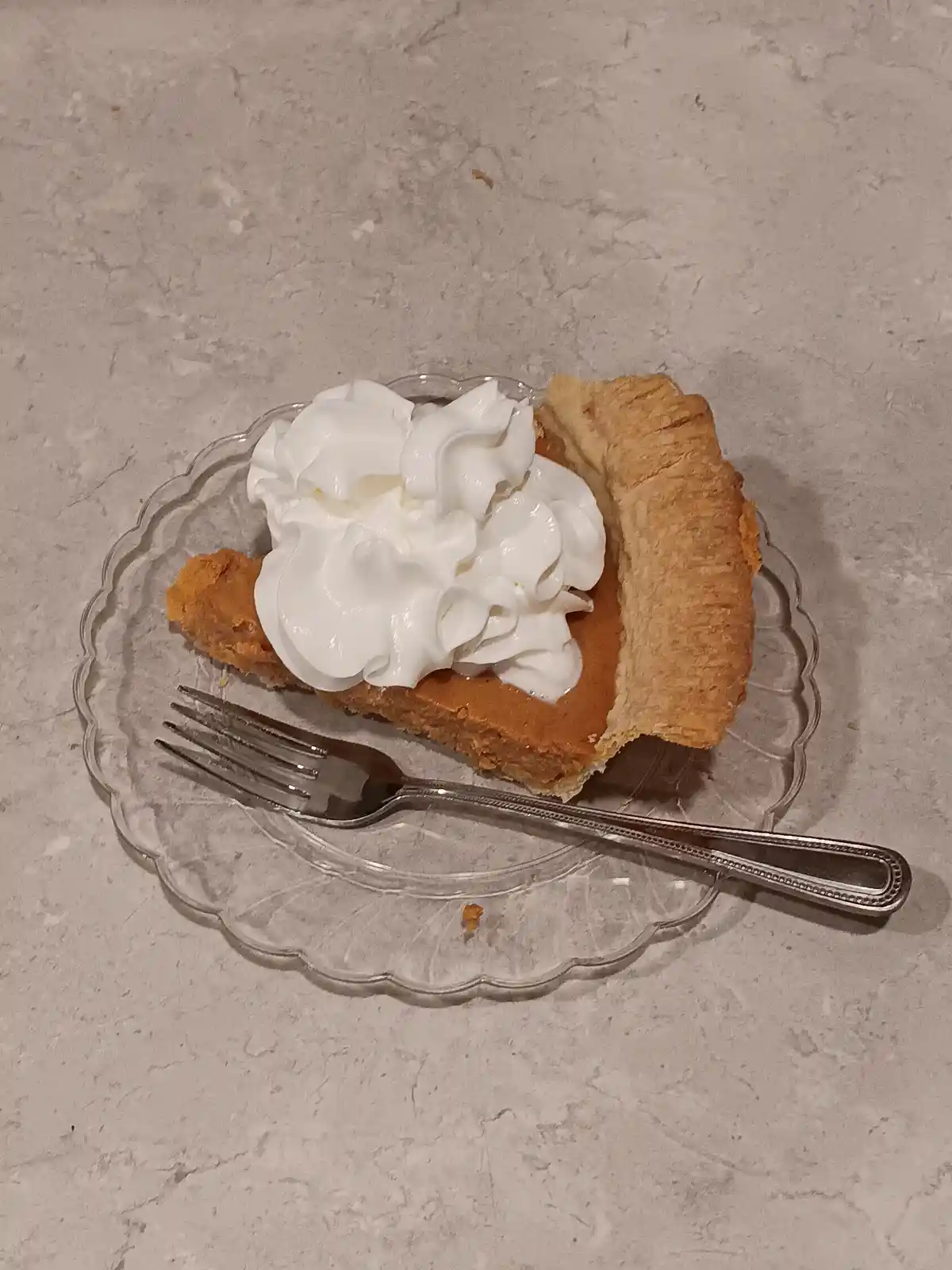 A slice of pumpkin pie with whipped cream sits on a clear glass plate with a fork beside it, implying the pie has been…