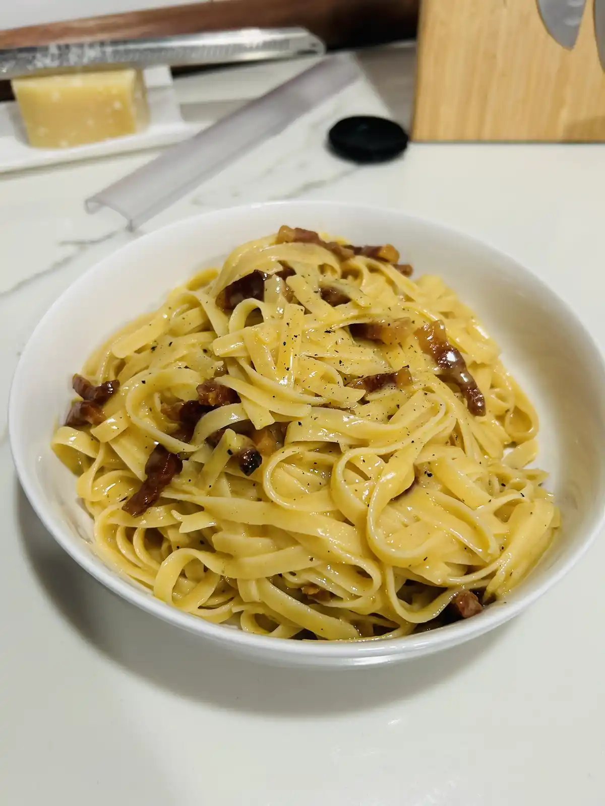 A close-up photo shows a white bowl filled with creamy fettuccine pasta mixed with crispy bacon bits and freshly ground…