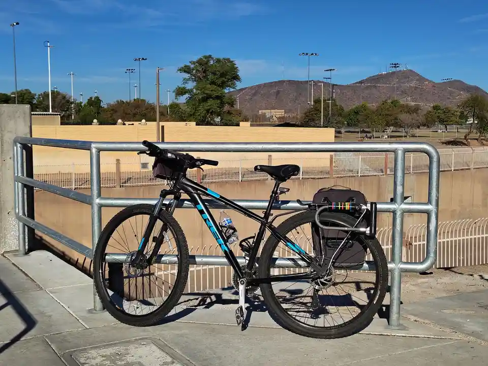 A black mountain bike with blue accents is parked against a metal railing on a sunny day, with a water bottle in a holder…