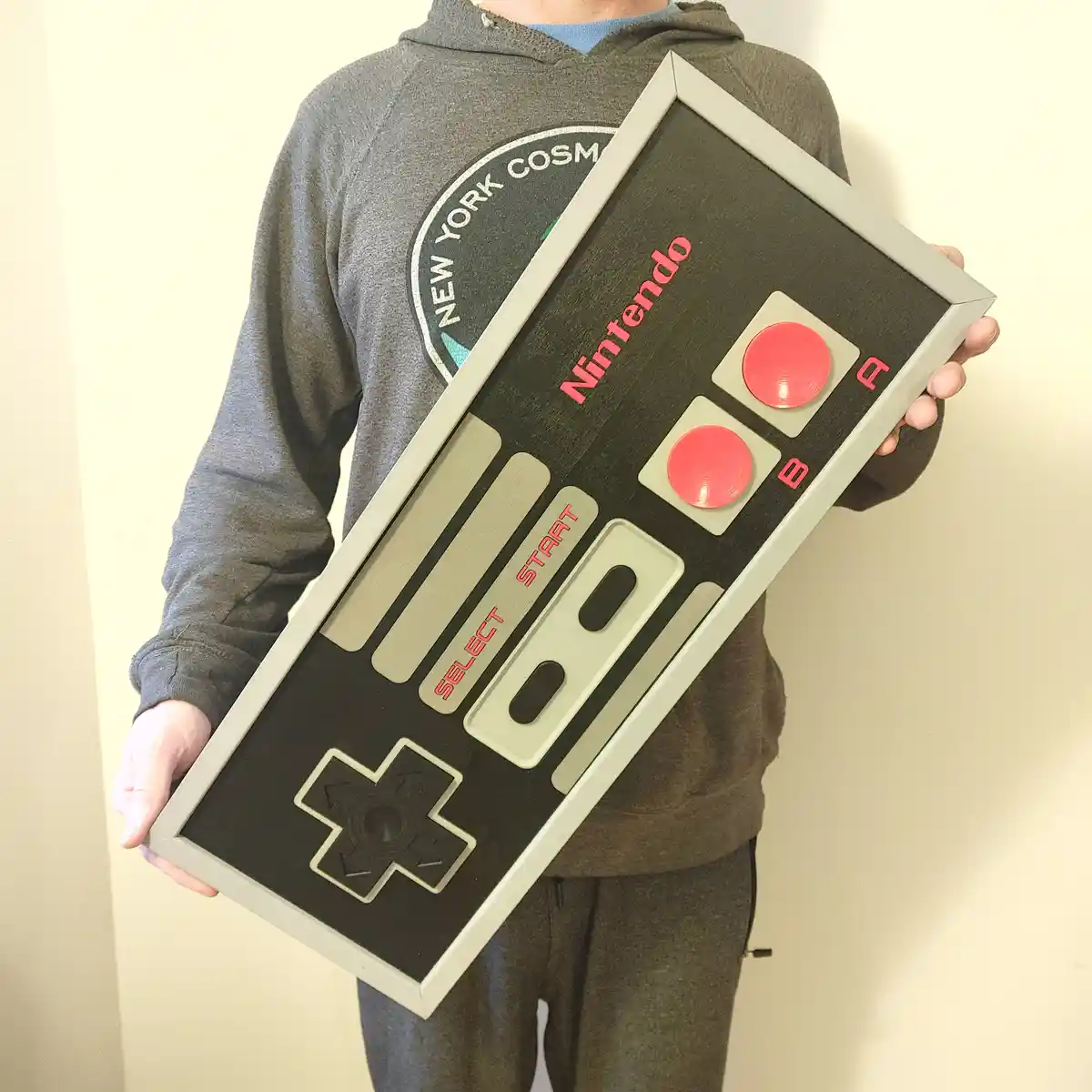 A person wearing a grey hoodie with a 'New York Cosmos' logo holds a large, framed replica of a classic Nintendo…