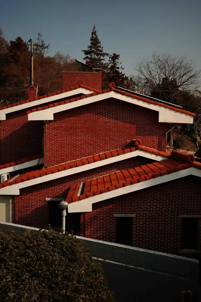A photograph of two red brick buildings with traditional tiled roofs under a clear sky, with trees and a low wall in the…