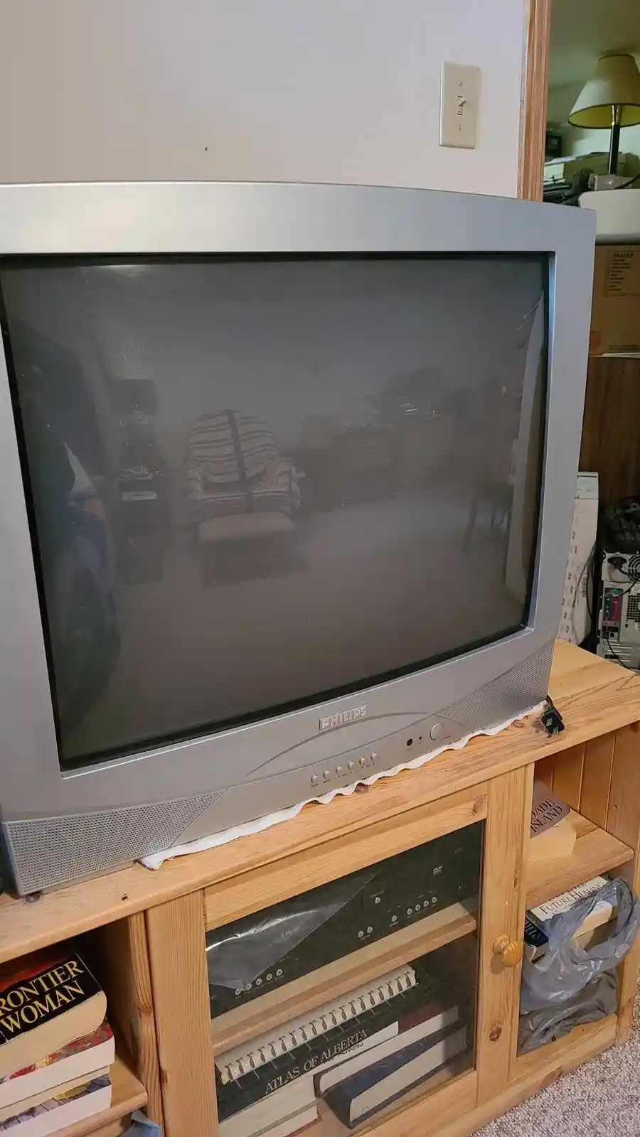 A silver Philips CRT television sits on a wooden media console, its screen dark and reflecting the room. Below it, books…