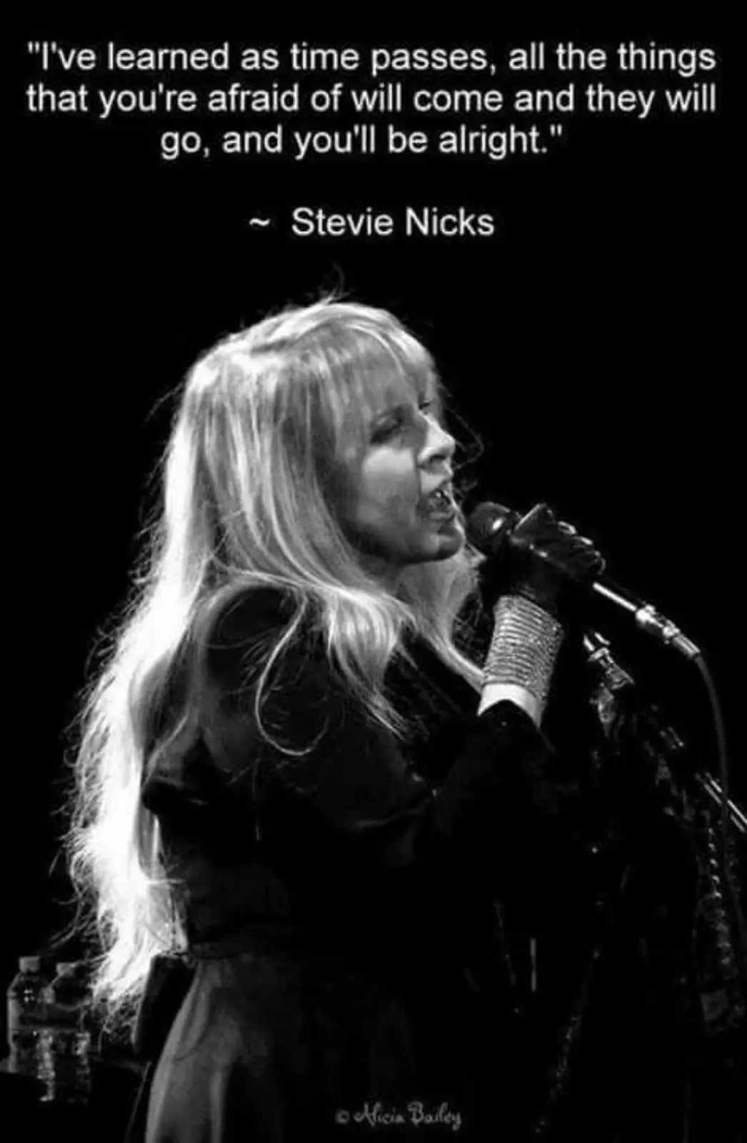 A black and white photo shows Stevie Nicks singing into a microphone on stage, her long blonde hair flowing. The image is…