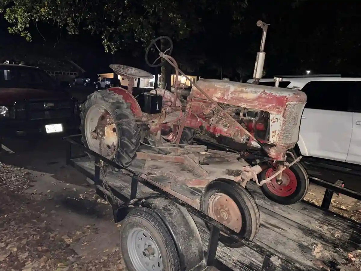 A rusted, red vintage tractor sits on a wooden flatbed trailer at night, surrounded by trees and parked cars. The tractor is…